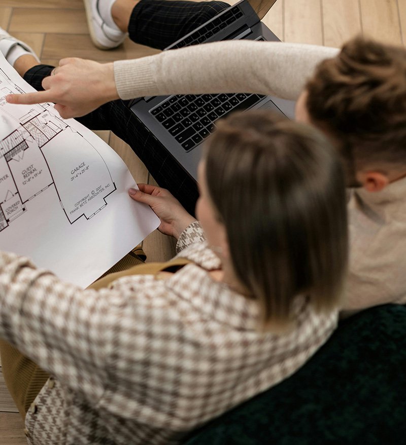 Dos personas sentadas consultando un plano y un ordenador portátil, vistos desde arriba