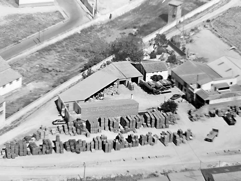 Vista aérea de instalaciones de empresa de construcción, con un par de naves y materiales apilados en el exterior. Foto antigua en blanco y negro.