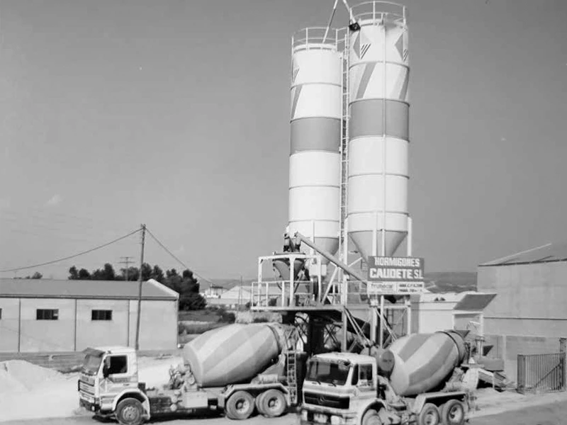 Planta de hormigón con dos camiones hormigonera cargando material. Foto en blanco y negro.